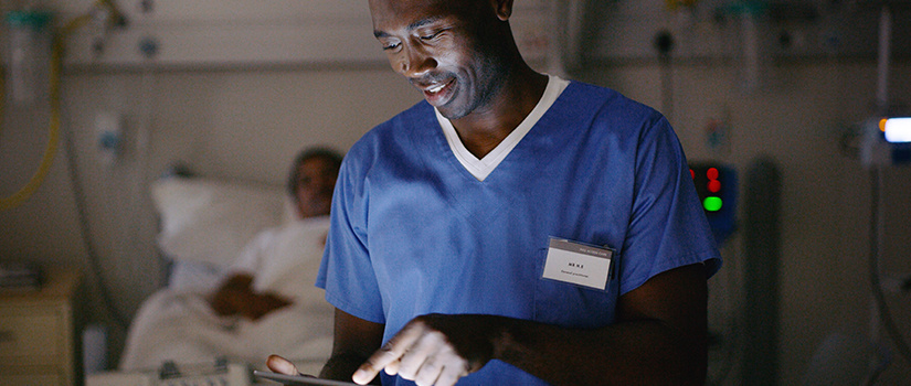 Image of a health care worker in a hospital pointing to an item on an iPad. A patient in a bed is seen in the background.