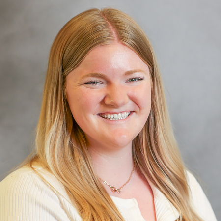 Professional headshot of a smiling person with long blonde hair wearing a white short-sleeve ribbed blouse and a delicate necklace. The person is facing the camera against a soft gray studio background with soft, even lighting, creating a friendly, approachable, and professional appearance.