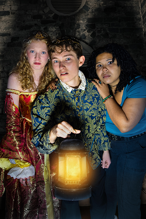 Three actors, tightly gathered together, looking into the camera at a unknown object. The center actor is holding a lantern with a ghostly yellow glow.