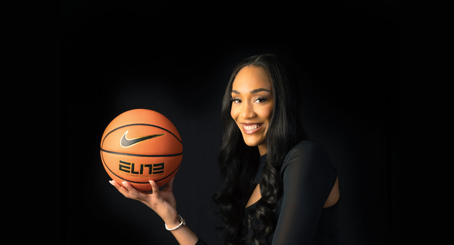 A woman with long black hair wearing a black sleeveless top and a delicate bracelet smiles brightly while holding a Nike Elite basketball in her outstretched hand. She is photographed against a dark black background in a professional portrait style."