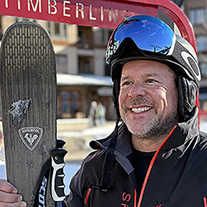 Man in ski helmet and goggles smiles while holding skis beneath a red Timberline sign at snowy mountain resort. 
